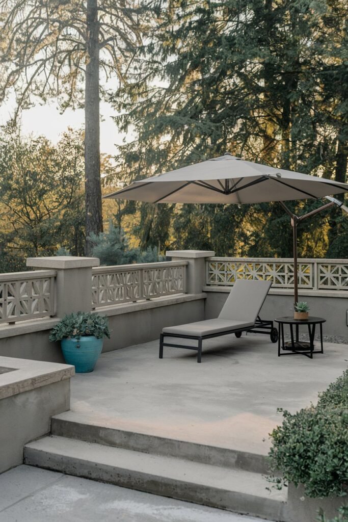 a backyard patio with a modern design. There's a lounge chair with an umbrella and a small table near the wall. A potted plant is placed on the table. The patio has a concrete floor and is surrounded by a low wall with a decorative railing. The background contains tall trees.