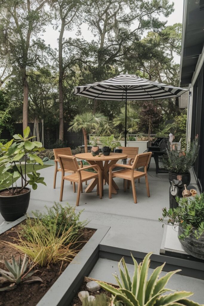 A backyard patio with a modern design. There is a table with four chairs made of teak wood. There are potted plants around the patio. The floor is made of gray concrete. A black and white striped umbrella is placed near the table. There are some gardening tools near the plants. The background contains tall trees.