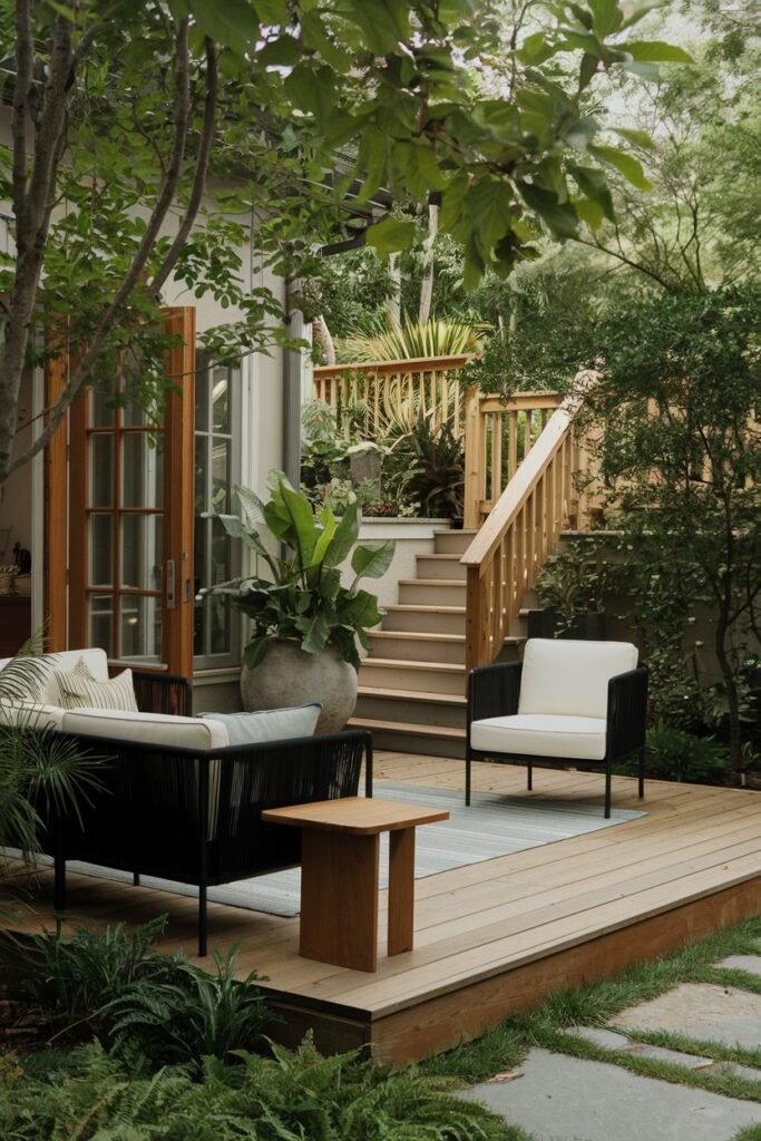 A backyard patio with a wooden deck, surrounded by lush greenery. There's a black outdoor couch and a white outdoor chair. A wooden side table is placed near the couch. A large potted plant is placed near the entrance. The background reveals a wooden railing and a set of stairs.