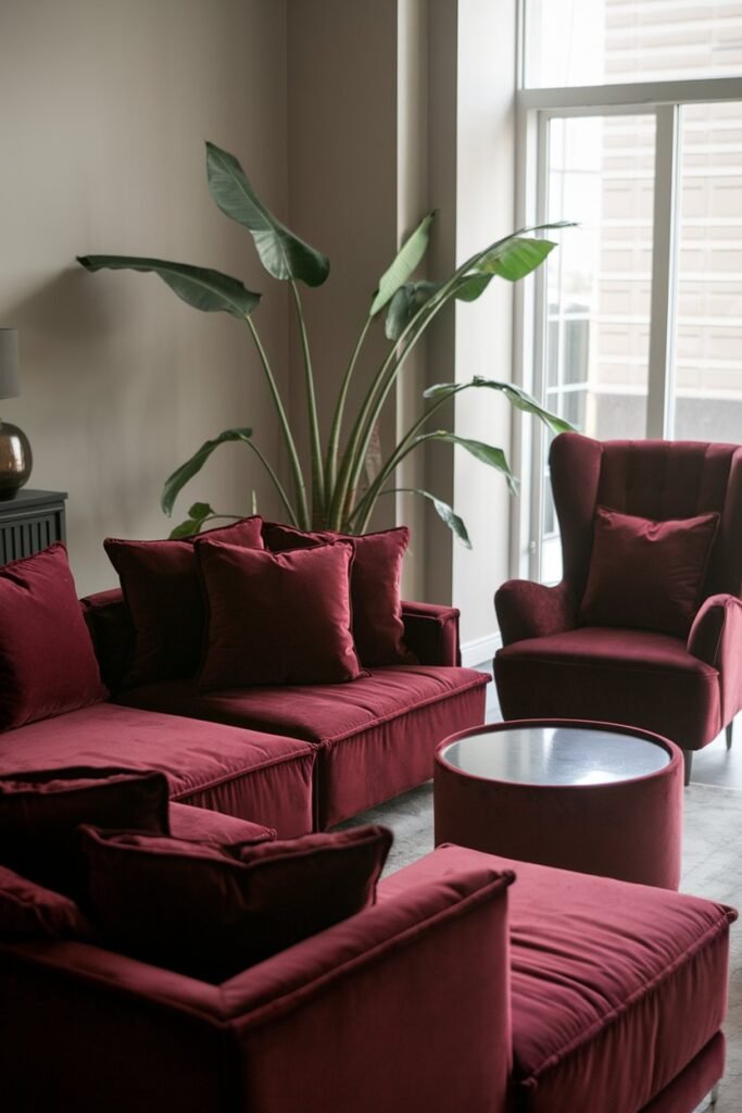 A cozy and elegant living room with a burgundy theme. The room has a large burgundy sectional sofa with multiple cushions. There's a matching burgundy armchair near the window. A small round burgundy coffee table is placed in front of the sectional sofa. A large potted plant with green leaves adds a touch of nature to the room. The walls are painted in a light gray color.