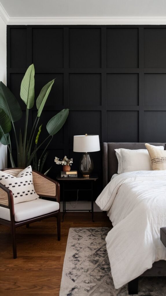A bedroom with a black accent wall. There's a bed with a white duvet and a few pillows. A nightstand with a lamp is placed next to the bed. A chair with a white cushion is placed in the corner. A large plant is placed near the black wall. The floor is wooden.