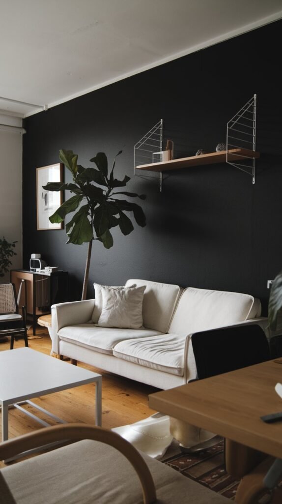 A living room with a black accent wall. There's a white sofa with a few pillows. A white coffee table is placed in front of the sofa. A potted plant stands next to the sofa. A wooden shelf is mounted on the black wall. There are a few decorative items on the shelf. The room has a few other pieces of furniture, including a chair and a table. The floor is made of wood.