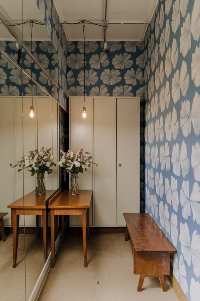 A small hall with a mirror wall. There is a wooden table with a vase of flowers. There is a storage unit with doors. There are hanging lights above the table. There is a wooden bench next to the wall. The wallpaper has a pattern of large white flowers on a blue background.