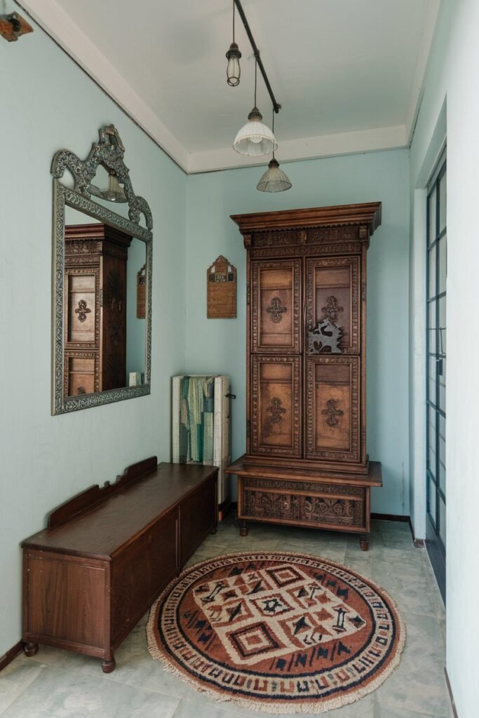 A small hall with a decorative mirror on the wall. There is a wooden storage cabinet with intricate carvings. Next to it, there's a wooden bench. On the floor, there's a round rug with a geometric pattern. Above the bench, there are three hanging lamps. The walls and the ceiling are painted in a light blue shade.