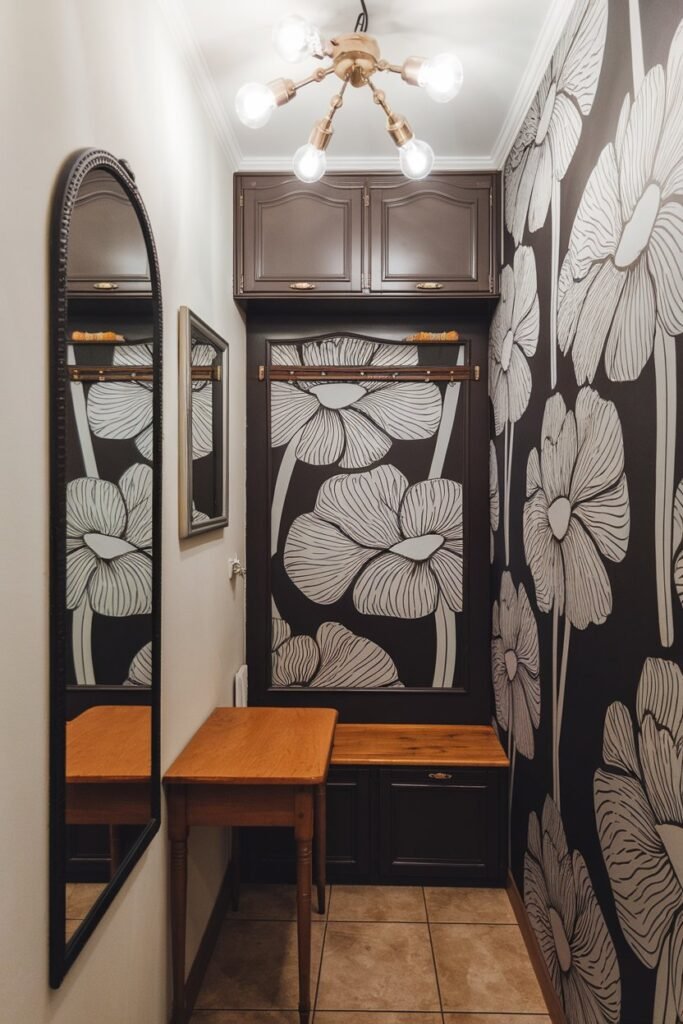 A small hallway with a mirror, a table, storage, lights, a bench, and wallpaper. The wall has a unique wallpaper with a large flower pattern. There is a wooden bench against the wall. Above the bench, there is a shelf with a vase. The floor is tiled. The hallway has a chandelier with multiple light bulbs.