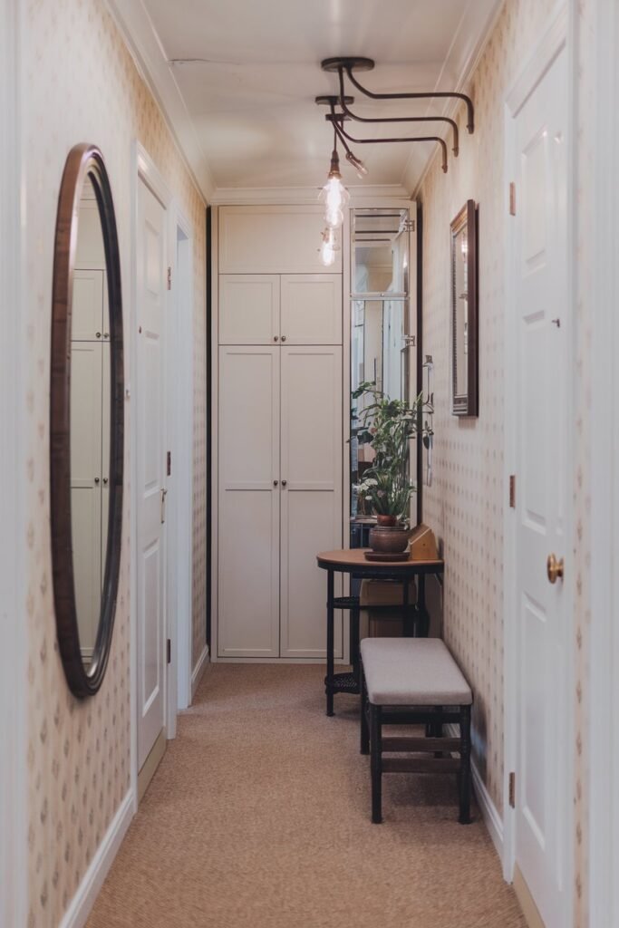 A small hall with a mirror, a table, storage, lights, a bench, and wallpaper. The hall has a beige carpet and white walls. The mirror is on the wall near the entrance. There is a small table near the mirror with a plant pot. There is a storage cabinet with doors near the table. The lights are hanging above the table. There is a bench near the table. The wall behind the bench has a patterned wallpaper.