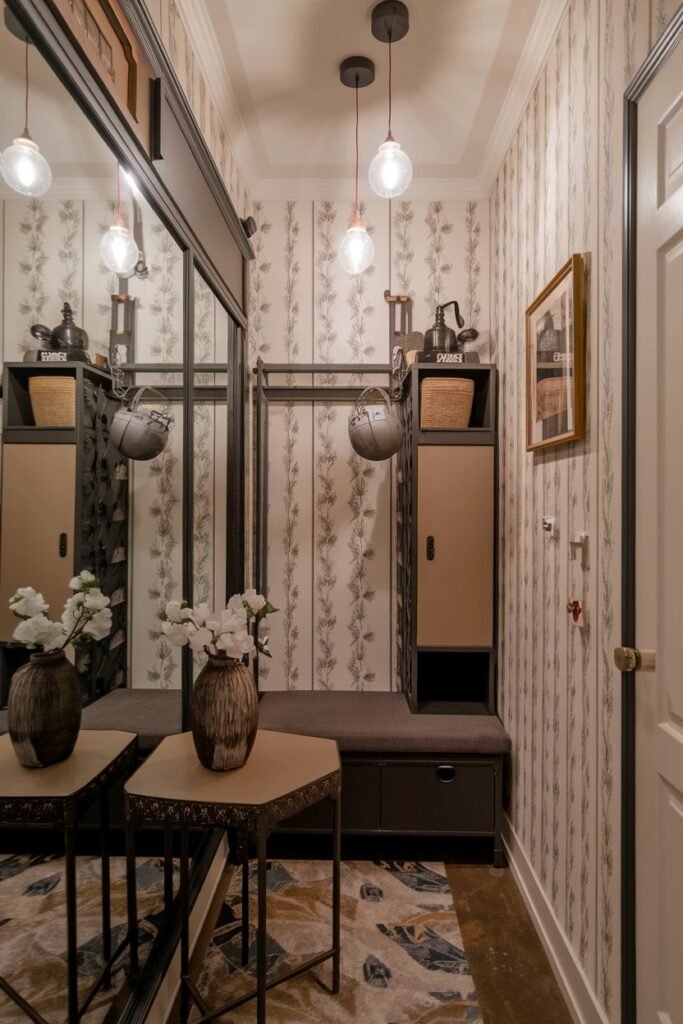 A small hall with a mirror wall. There's a table with a vase and a candle. There's a bench against the mirror wall and a storage cabinet. The room has hanging lights. The wallpaper has a floral pattern.