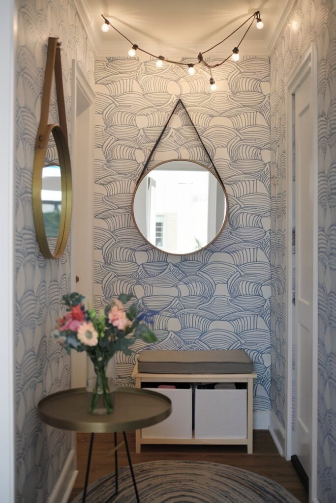 a small hallway with decor ideas. There is a round mirror hanging on the wall. On the floor, there is a round table with a vase of flowers. Against the wall, there is a storage bench with a cushion. Suspended from the ceiling, there are string lights. The wallpaper has a pattern of blue and white waves.