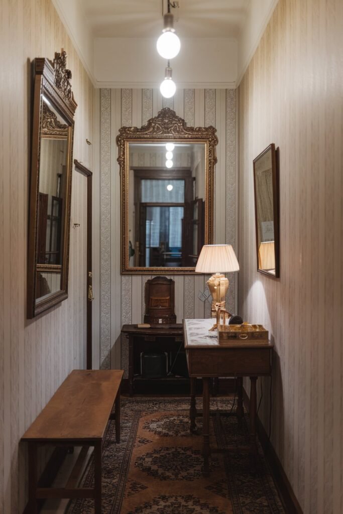 A small hallway with a mirror on the wall. There is a wooden table with a lamp and a few items on it. There is a wooden bench against the wall. The floor is covered with a rug. The walls have a patterned wallpaper. There are lights hanging from the ceiling.