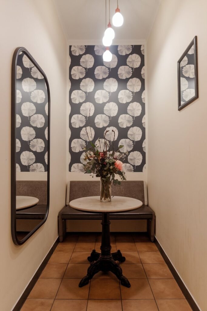 A small hall with a mirror on one wall. There's a round table in the center with a vase of flowers. On the other side of the room, there's a bench against the wall. Above the table, there are hanging lights. The wall opposite the mirror has a wallpaper with a pattern of large white flowers on a dark background. The floor is tiled.