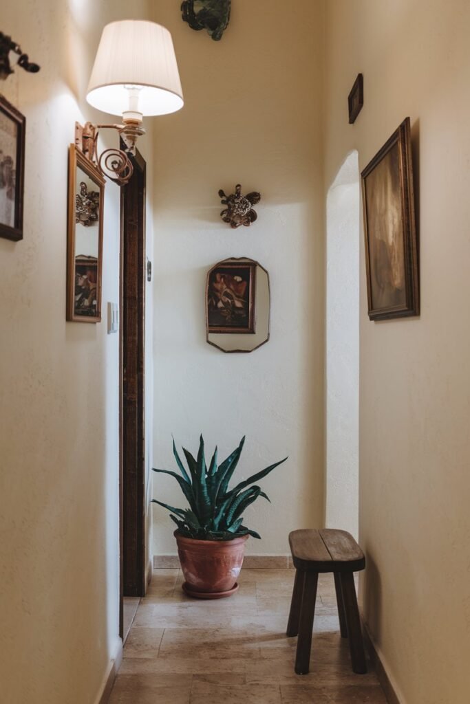 a small hallway with a few items. There is a lamp on the left wall, a wooden stool next to the right wall, and a plant in a pot on the floor. The walls have a beige color and a few decorations, including a mirror and a painting. The floor is made of beige tiles.