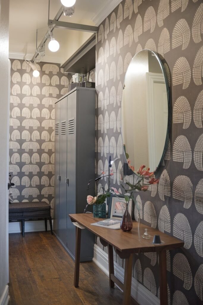 a small hall with a mirror, a wooden table, a storage unit, lights, a bench, and a unique wallpaper. The wallpaper has a pattern of large, white, geometric shapes on a dark grey background. The mirror is hung on the wall above the wooden table. The wooden table has a few items on it, including a vase with flowers and a lamp. The storage unit has doors and is placed near the wall. There are lights hanging from the ceiling. The flooring is hardwood.