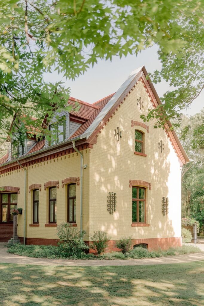 An outdoor shot of a yellow brick building with red trim, dark windows, and a red roof. There are trees surrounding the house, as well as a green lawn.