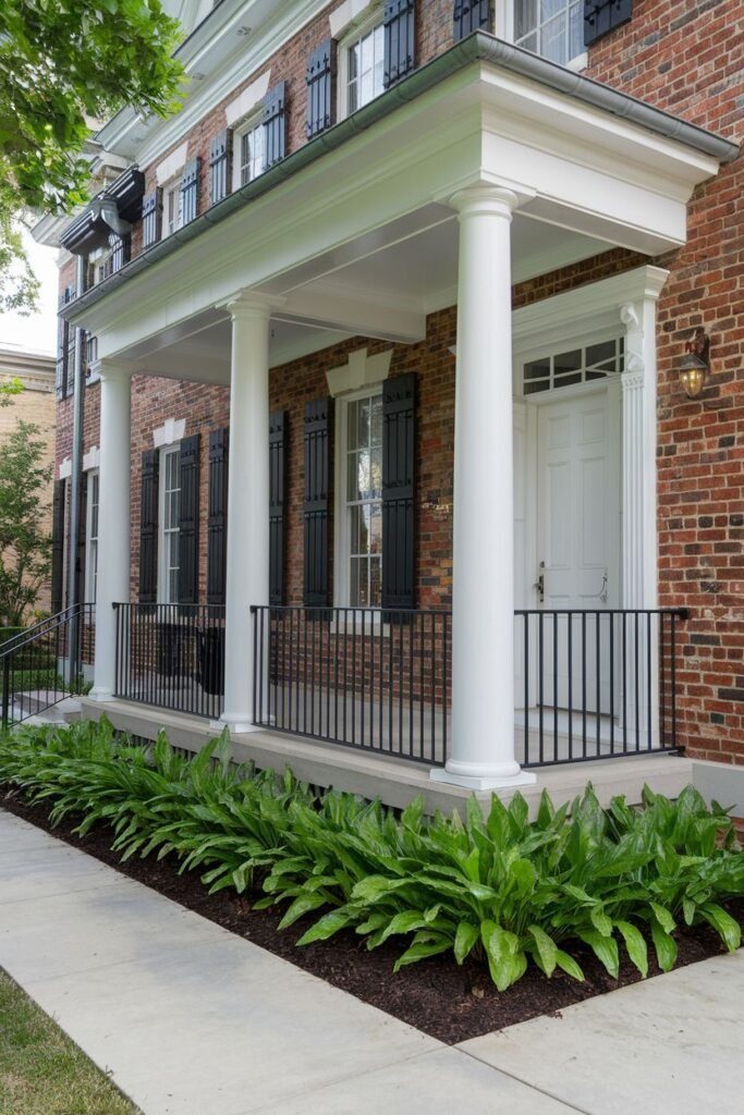 A close, medium outdoor shot of a red brick house with white columns, a black railing and black shutters. The foreground is landscaped with green plants and a sidewalk.