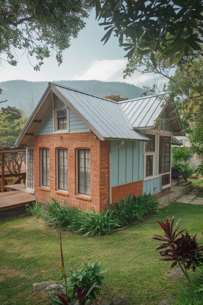 A medium outdoor shot of a small, light-blue house with a brick lower half and a silver roof. There is a green lawn in front of the house with various plants, and the background shows mountains and trees.