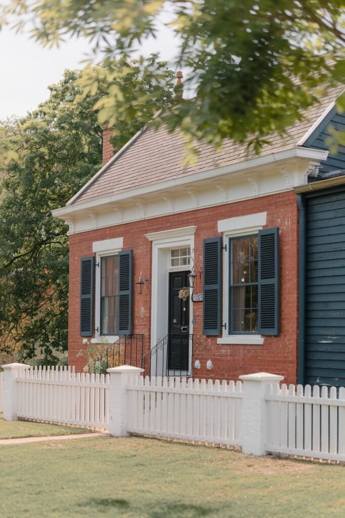 A straight on shot of a red brick house with black shutters and a black door. The house is situated behind a white picket fence. There are trees surrounding the house.