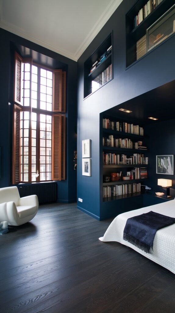 A bedroom with dark blue walls and built-in bookshelves, a large window with wooden shutters, a dark hardwood floor and a white armchair.