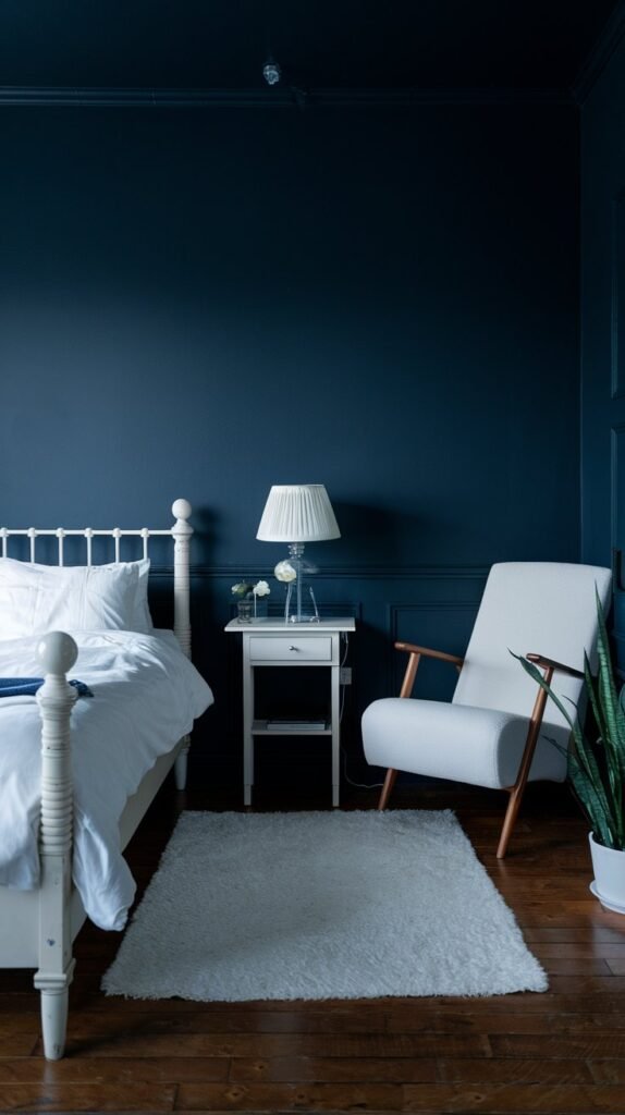 A simple bedroom with dark blue walls, dark hardwood floors, a white bed, a white chair, and a fluffy white rug.