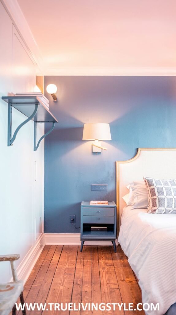 A moody yet stylish bedroom with blue walls and warm wooden floors. A bed with a white headboard and striped bedding is shown, along with a small blue nightstand and wall-mounted lighting.