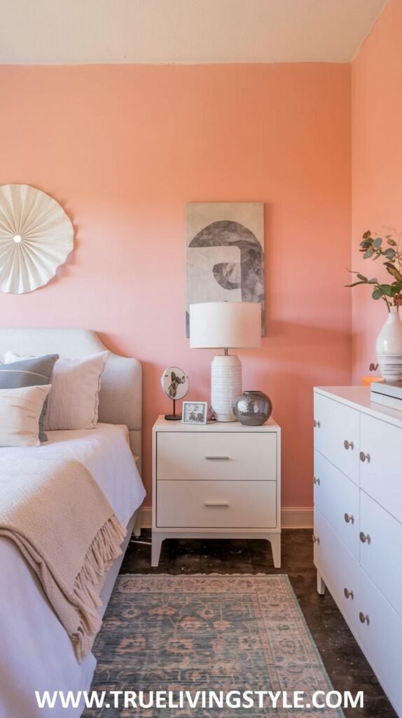 A warm bedroom with soft coral walls and white trim. A bed with a light upholstered headboard and white bedding with a blush-colored throw is featured, along with a white dresser.