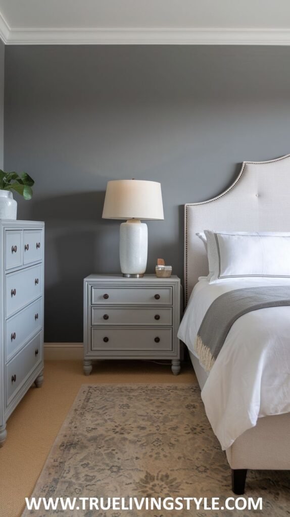 A sophisticated bedroom with dark gray walls and white trim. A bed with a light upholstered headboard and white bedding with a gray throw is visible, along with a light blue dresser and nightstand.