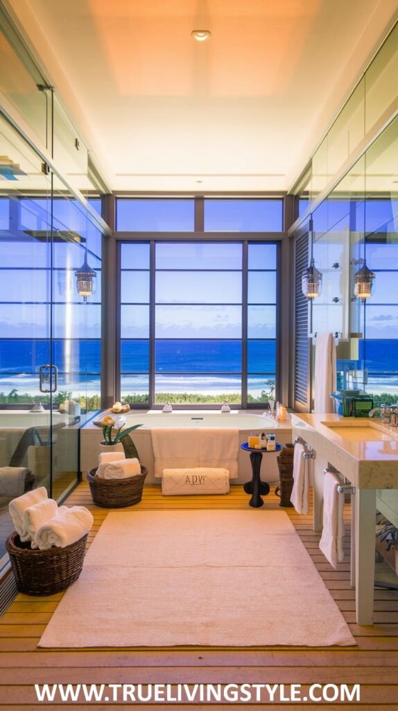 An open and airy coastal bathroom includes a large window, glass shower enclosure, and light wood flooring, complemented by plants and woven baskets.