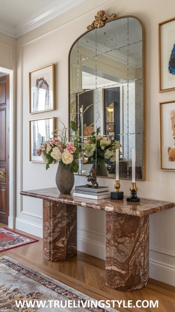 A marble-topped console table displays a vase of flowers, books, and candlesticks, in front of a large multi-paneled mirror with framed artwork on the surrounding wall.