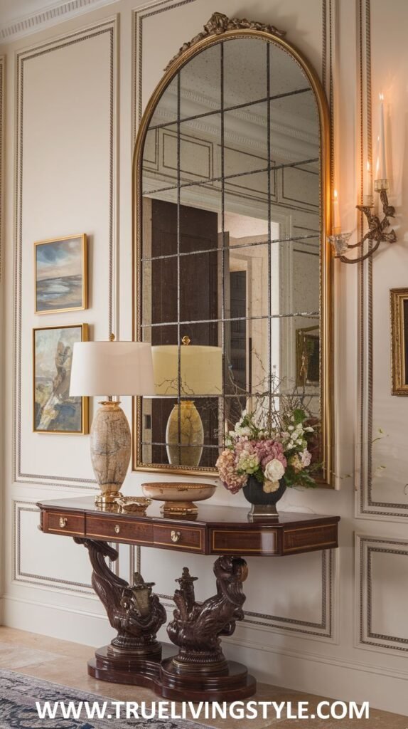 A wooden console table with drawers supports two lamps, a decorative bowl, and a vase of flowers, set against a panelled wall with a large arched mirror and wall sconces.