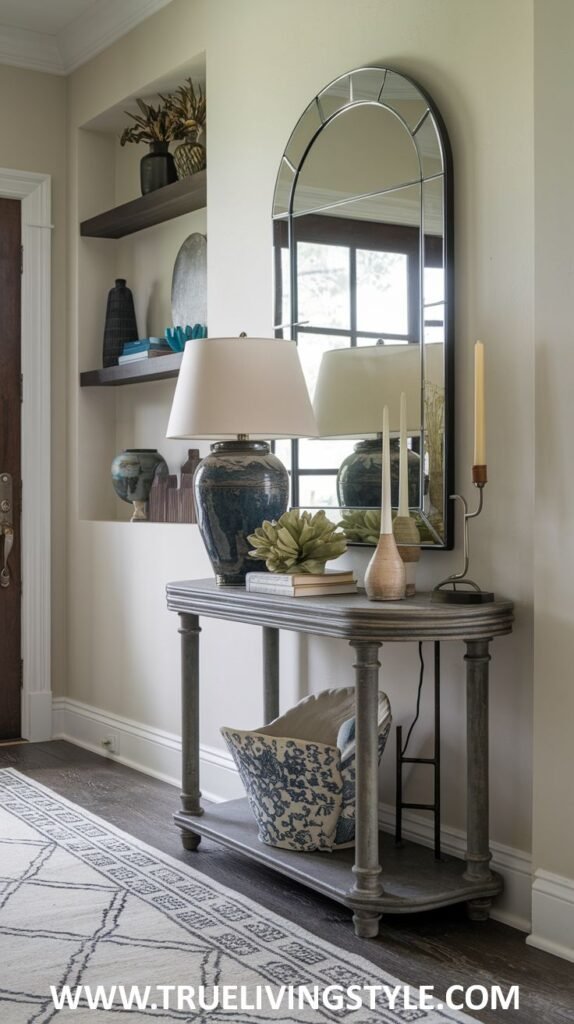 A grey console table with a lamp, vases, and candlesticks has a patterned basket underneath, placed before an arched mirror with decorative shelves visible in the reflection.