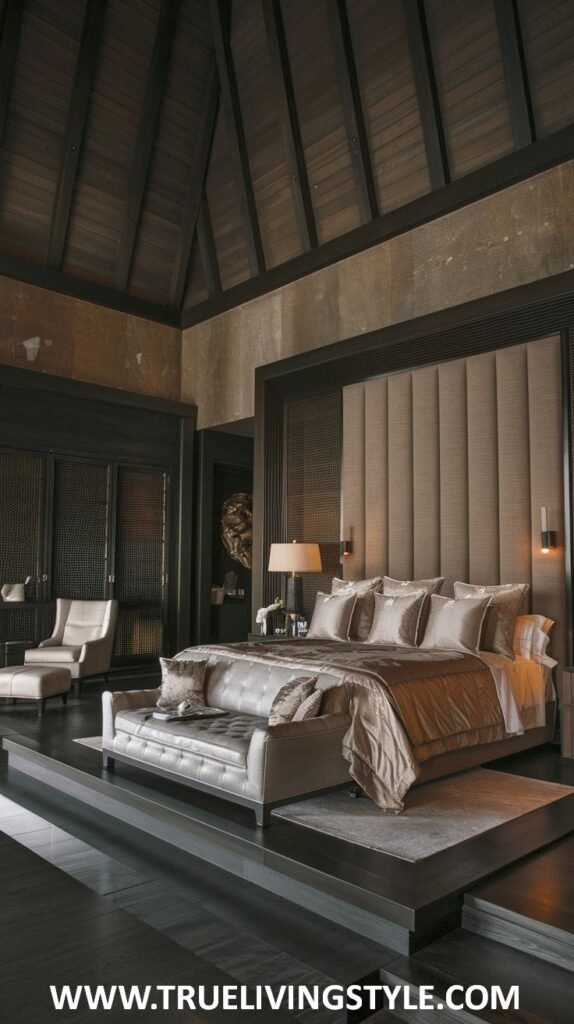 A dark earthy bedroom with dark wood accents and a high ceiling features a raised platform bed with light beige bedding and a gold throw, a matching upholstered bench, a light armchair, and dark wood flooring.