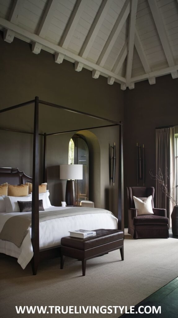A dark earthy bedroom features a four-poster bed with olive green canopy, white bedding, a brown upholstered bench at the foot of the bed, a brown armchair with a white pillow, and dark brown walls under a white beamed ceiling.