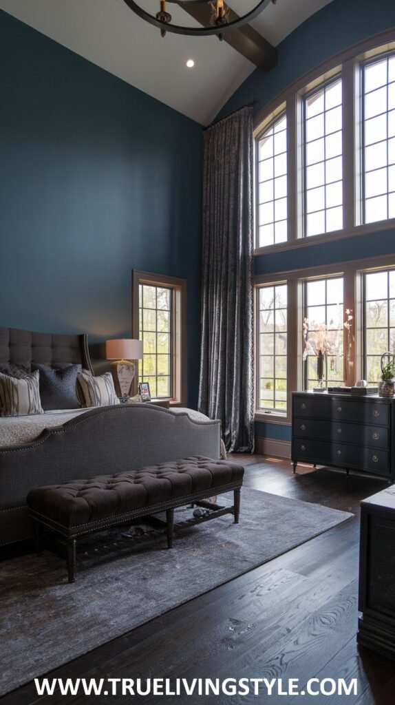 A dark earthy bedroom with dark blue walls and high ceilings features a large upholstered bed with patterned pillows, a dark brown tufted bench, a dark wood dresser under large windows with patterned curtains, and dark wood flooring.