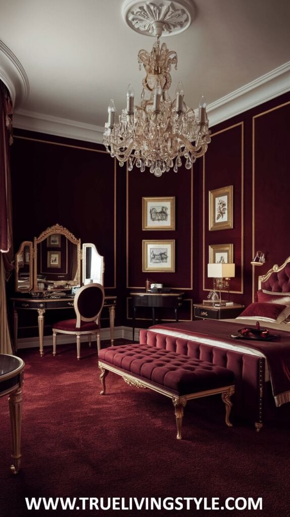 A luxurious dark red bedroom features a striking crystal chandelier hanging from the ceiling, classic dark-toned furniture with gold accents including a vanity table and chair, and a plush dark red upholstered bench at the foot of a bed partially visible on the right. Framed traditional artwork adorns the dark red walls.