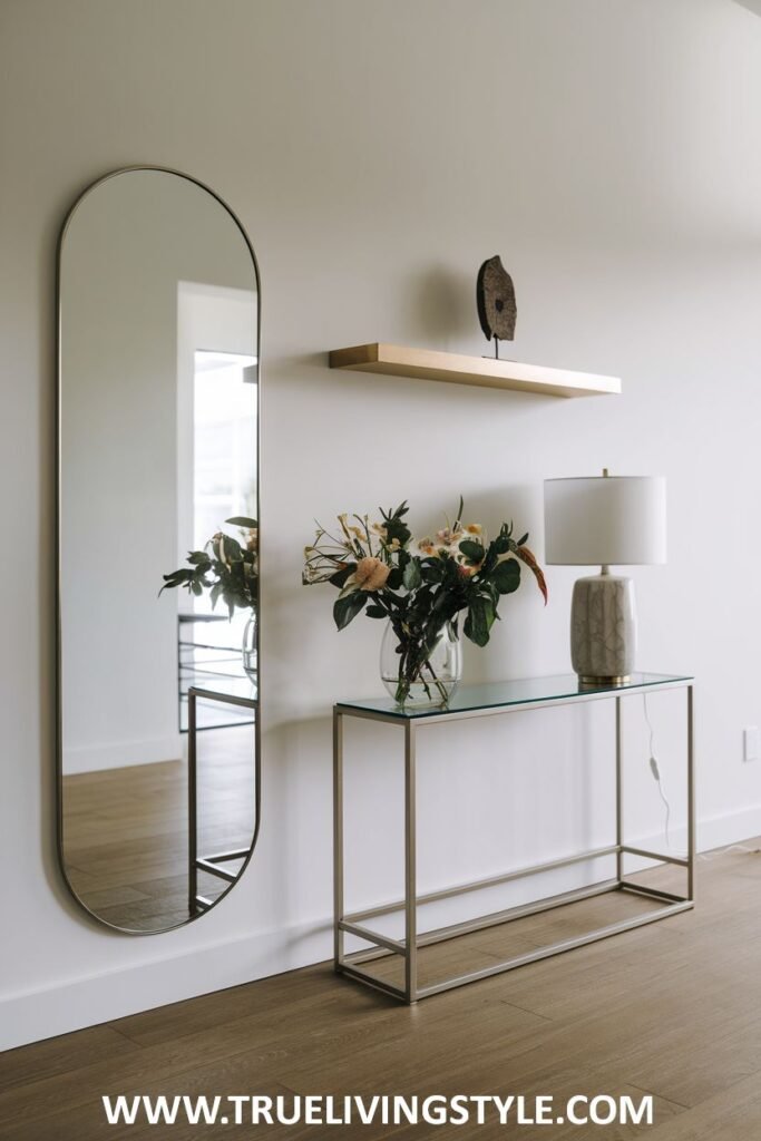 A tall mirror next to a glass console table with flowers and a lamp.