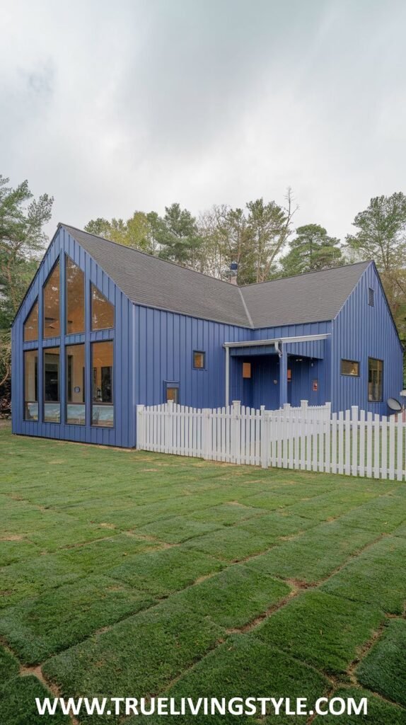 A modern barn-style house painted in a bold blue, with tall, prominent windows, a dark roof, and a white picket fence surrounding a green lawn.