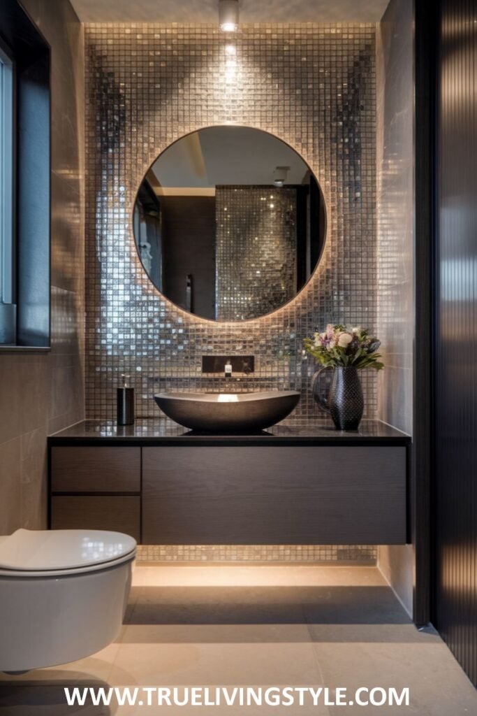 A stylish luxury powder room with a wall covered in shimmering mosaic tiles, a round mirror with integrated backlighting above a dark countertop with a vessel sink, and subtle under-cabinet lighting. A toilet is part of the space.