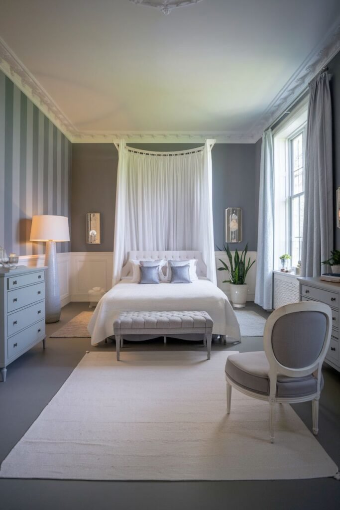 This is a grey and white bedroom with warm lighting. A four-poster bed with white sheer fabric, a light-colored rug, and a grey armchair are the main elements.