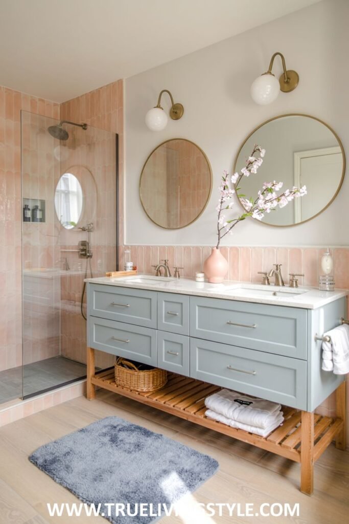 A pastel blue and peach bathroom includes a glass-enclosed shower, a light blue double vanity with a light countertop, round gold mirrors, and light wood flooring. This is a view into a pastel bathroom.