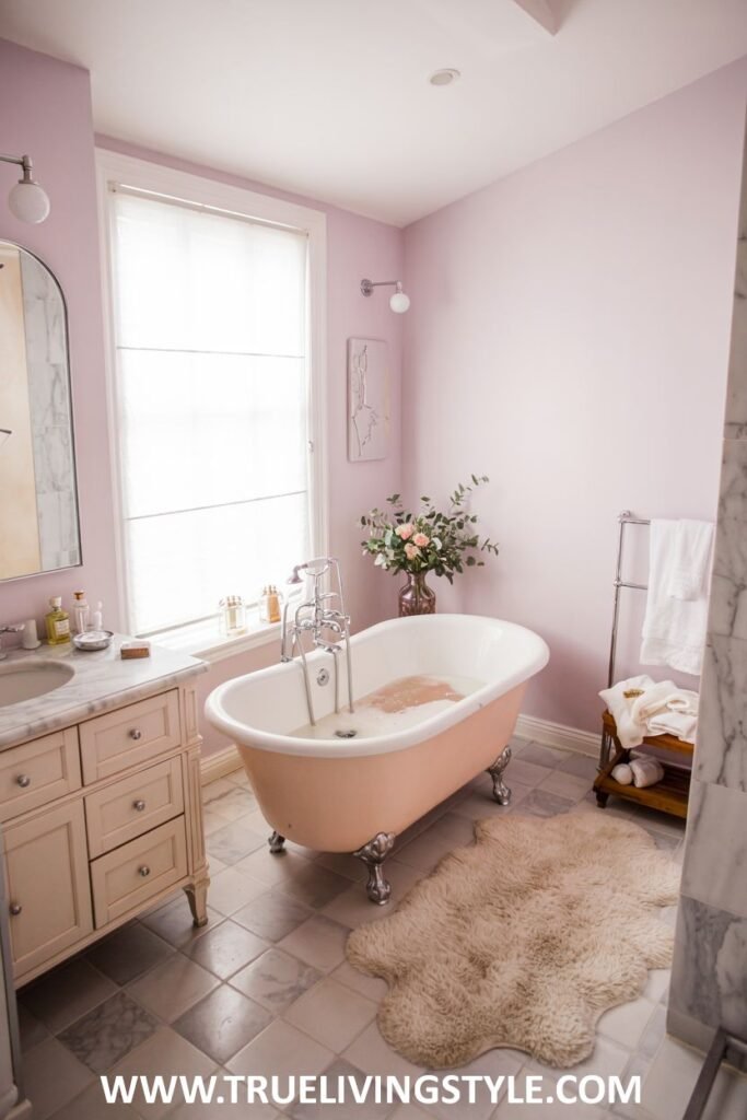 A pastel lilac bathroom features a light pink clawfoot bathtub, a light wood vanity with a marble top and an arched mirror above, and patterned floor tiles. This is a pastel bathroom.