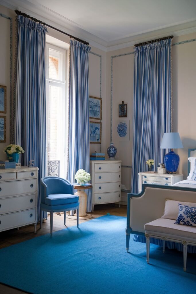 A vibrant comfortable french bedroom decorated with bold blue and white striped curtains and a bright blue rug. The room contains white dressers, a blue armchair, and a bench.