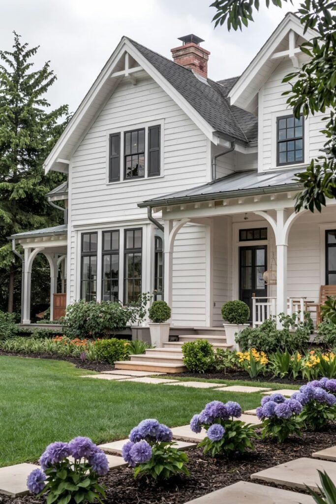 A close-up view of a white farmhouse with a large porch, dark framed windows, and extensive landscaping featuring purple and yellow flowers along a stone pathway.