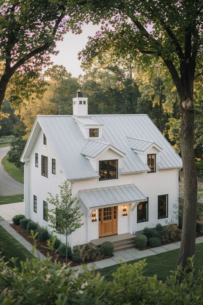 An aerial-style view of a white farmhouse with a metal roof and multiple dormer windows. The house has light-colored double front doors and is surrounded by trees and landscaping.