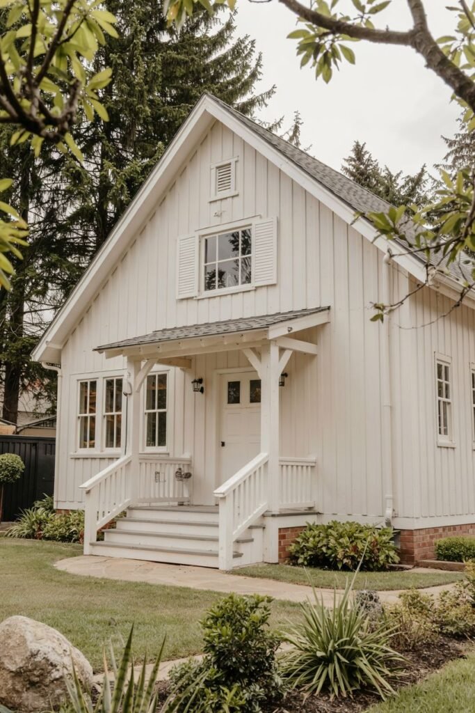 A small white farmhouse with a porch and a peaked roof. It has white shutters on the upper window and landscaping around the foundation.
