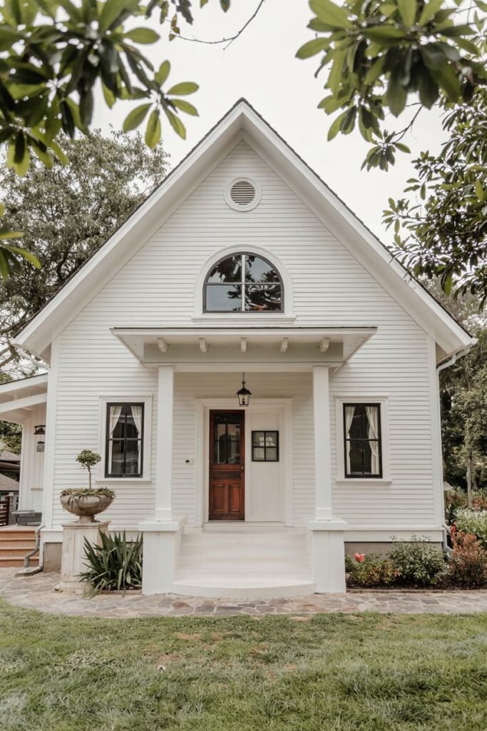 A white farmhouse with a dark wooden door and a large arched window above the porch. Landscaping is visible around the foundation.