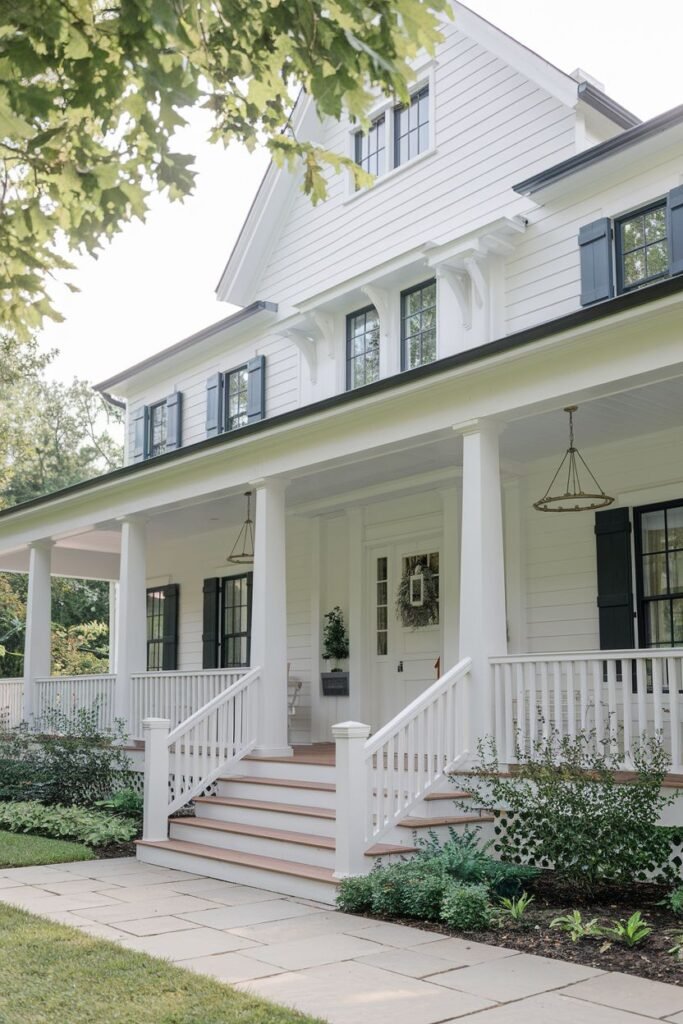 A large white farmhouse with a wide porch supported by white columns. White railings line the steps and porch. Dark shutters are on the windows of the second floor.